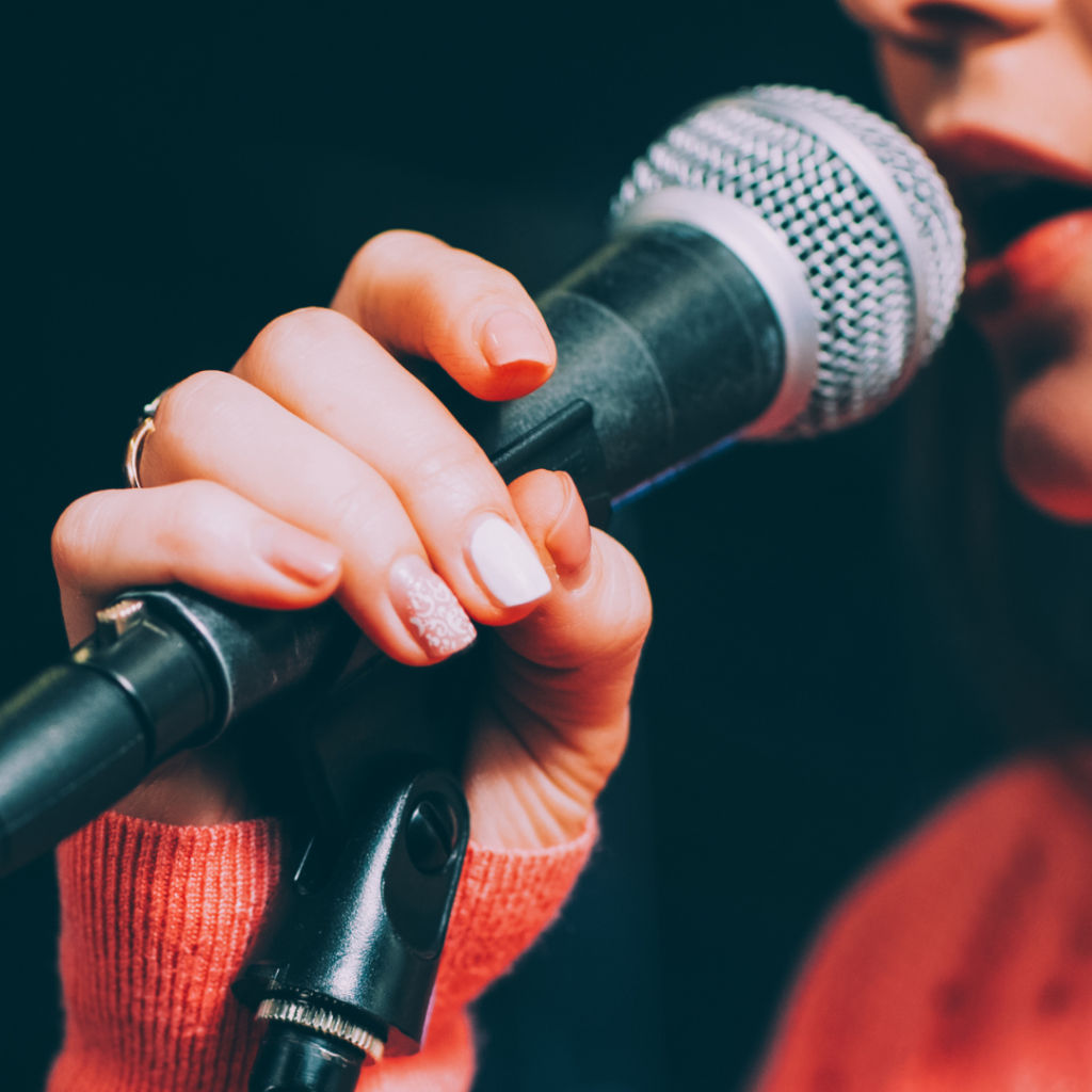 Abbildung zeigt die Hand sowie die Mundpartie einer Frau, die in ein Mikrofon singt