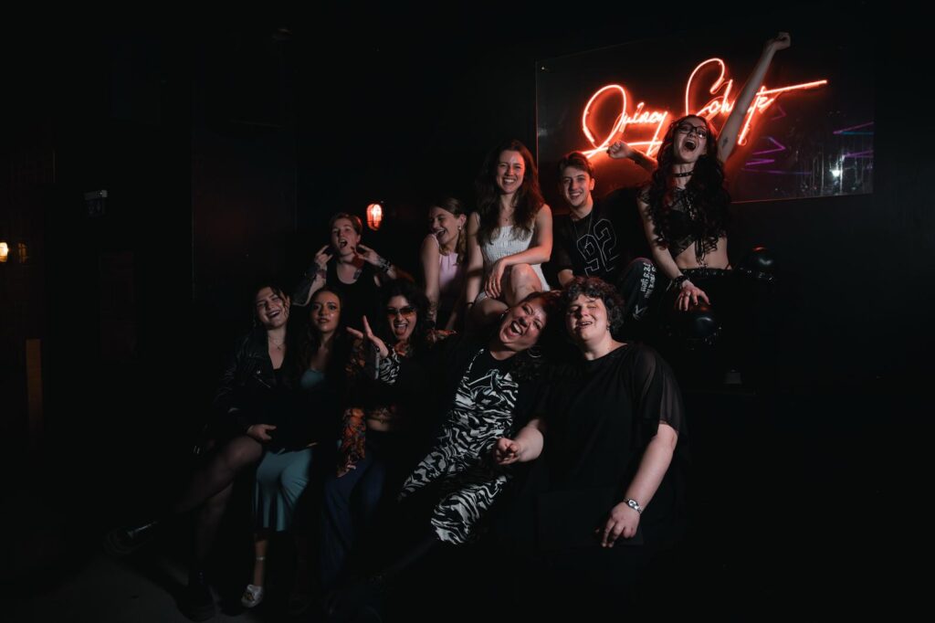 Elf Menschen in bunter Kleidung beim Abschlusskonzert meines Gesangsstudiums, vor einem leuchtenden „Queer Soft“-Neonschild – ein Moment voller Ausdruck und Verbundenheit